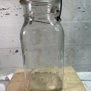 Op de afbeelding: Een glazen pot met een draadbeugel sluiting en een rode rubberen afdichting. De pot is gelabeld met "Hazel-Atlas Glass 1/2 Cup".