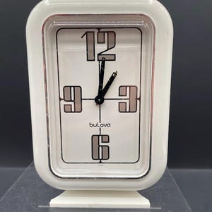 Puede incluir: Un reloj despertador Bulova vintage en una caja rectangular blanca. La esfera del reloj tiene manecillas negras y números grandes y estilizados en el 12, 3, 6 y 9. La marca "Bulova" está impresa en la esfera. El reloj está hecho en Japón.