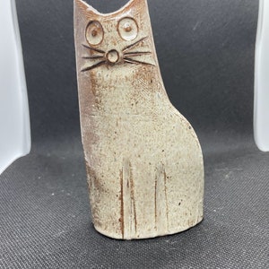 Puede incluir: Una figurita de cerámica de un gato con un esmalte blanco y marrón. El gato tiene ojos grandes y redondos y una cara simple y estilizada.