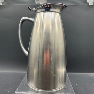 May include: A tall, brushed metal pitcher with a curved handle and a hinged lid. The pitcher has vertical ridges and a spout for pouring. It is displayed on a clear, rectangular stand.