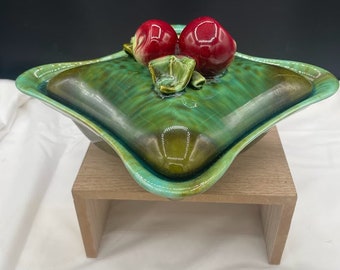 Puede incluir: Un plato para servir de cerámica verde y azul con tapa. La tapa tiene dos manzanas de cerámica roja en la parte superior. El plato está sobre un soporte de madera.
