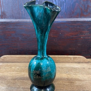 May include: A vintage, teal-colored ceramic vase with a flared, ruffled rim. The vase has a bulbous base and a slender neck. Gold lettering on the vase reads "Niagara Falls Canada". The vase is approximately 8 inches tall.