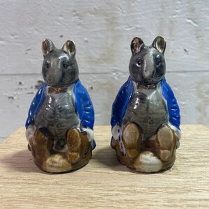 May include: A pair of ceramic salt and pepper shakers shaped like mice wearing blue jackets and brown pants. The mice are sitting on a wooden surface.