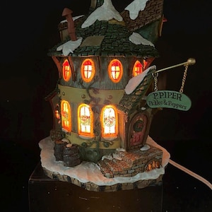 May include: A whimsical, illuminated ceramic house with a snow-covered roof and windows. The house features a sign that reads "P. PIPER Pickles & Peppers." The windows glow with warm light, and the house is set on a snowy base.