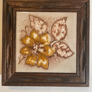 May include: A framed ceramic tile with a brown and yellow floral design. The tile is set in a dark wood frame.