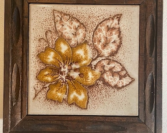May include: A framed ceramic tile with a brown and yellow floral design. The tile is set in a dark wood frame.