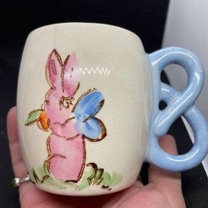 May include: A vintage ceramic mug with a light blue handle. The mug features a hand-painted design of a pink bunny holding a blue butterfly.