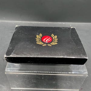 Op de afbeelding: Een zwarte doos met een rood en goud embleem op de bovenkant. Het embleem heeft een rode bol met een witte wervel, omringd door een gouden krans. De doos staat op een helder, reflecterend oppervlak.