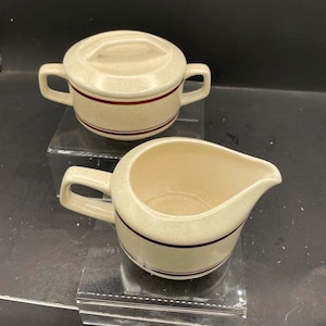 Op de afbeelding: Een crèmekleurige keramische suikerpot met deksel en twee zijhandvatten, gecombineerd met een bijpassende melkkan. Beide stukken hebben een gespikkelde afwerking en twee dunne, donkerbruine horizontale strepen rond de onderkant.