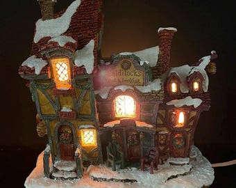 May include: A detailed, illuminated miniature house with a snow-covered roof and base. The house features multiple windows, a chimney, and the text "Goldilocks Bed & Breakfast". The house has a warm, inviting glow.
