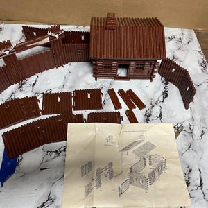 Puede incluir: Un juego de cabaña de juguete de plástico marrón con una cabaña, vallas y otros componentes. El juego incluye una cabaña con chimenea, vallas con elementos decorativos y un diagrama. Las piezas están dispuestas sobre una superficie estampada en blanco y gris.