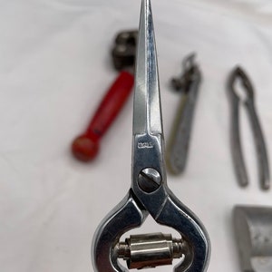 May include: Close-up of a silver pair of pruning shears with a pointed tip. The shears have a small metal roller in the middle of the handles. The word "ITALY" is engraved on the side of the shears.