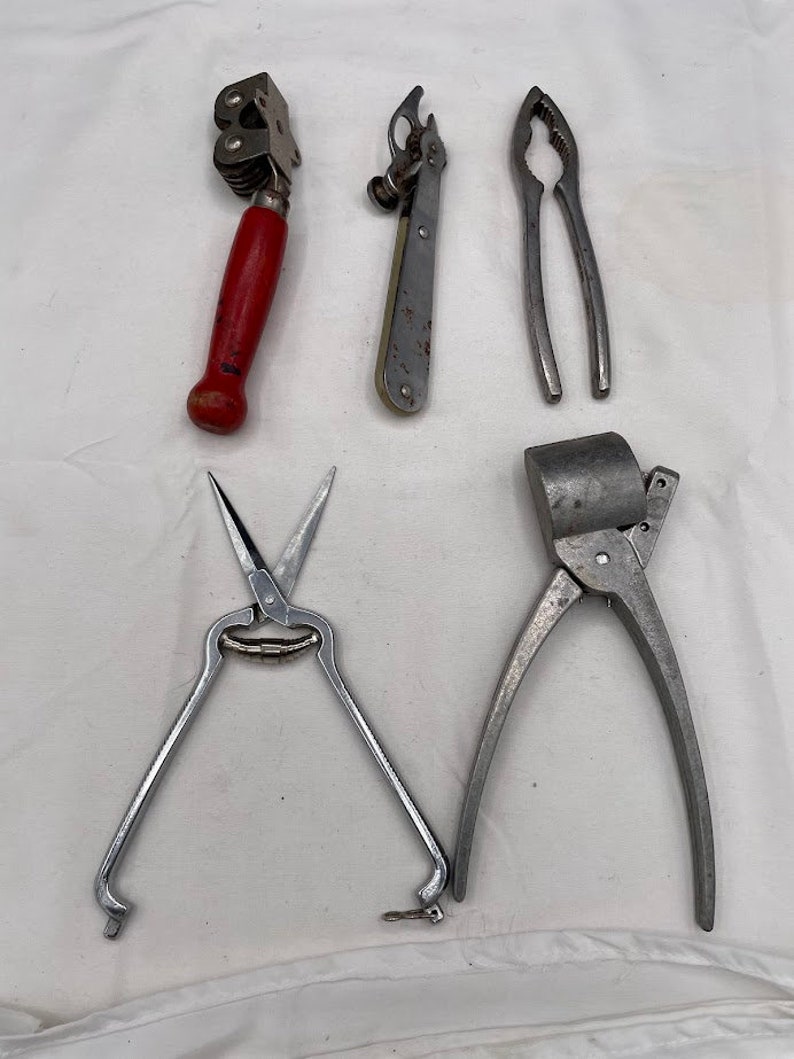 May include: Five kitchen tools: a red handled can opener, a silver nutcracker, a silver garlic press, a silver pair of kitchen shears, and a silver can opener.