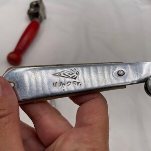 May include: A silver can opener with a red handle. The can opener has a diamond-shaped logo and the text "W. Nr. 25 K."