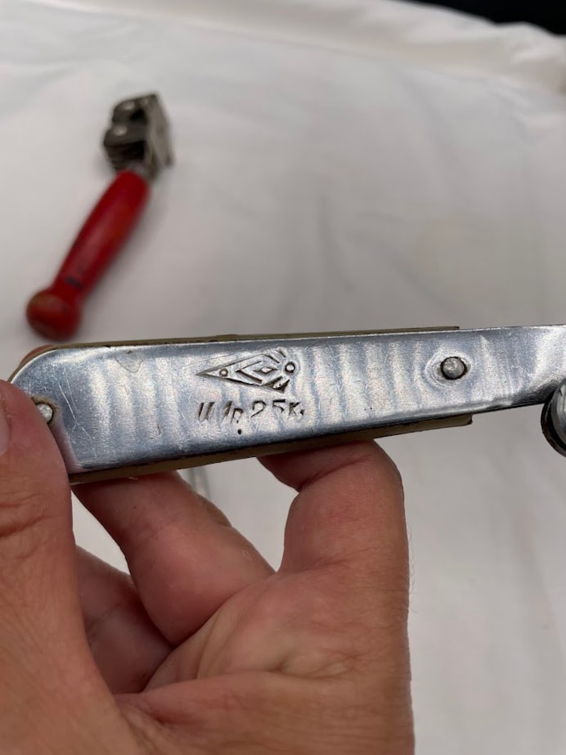 May include: A silver can opener with a red handle. The can opener has a diamond-shaped logo and the text "W. Nr. 25 K."