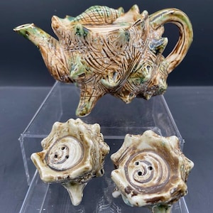 May include: A ceramic teapot and two matching salt and pepper shakers shaped like seashells. The teapot is brown and green with a textured surface. The salt and pepper shakers are white with brown accents.