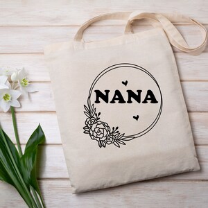 May include: A white tote bag with black text that reads "NANA" inside a circle with a floral design. There are three small hearts around the text.