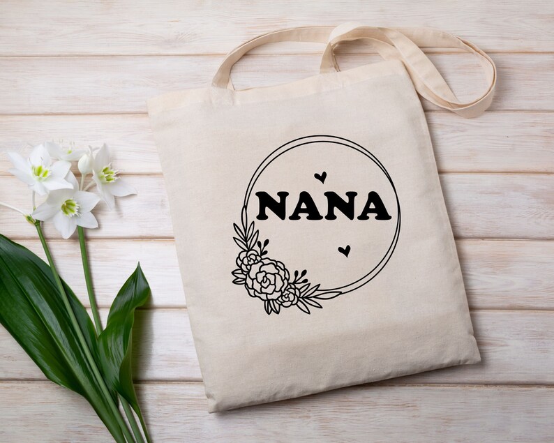 May include: A white tote bag with black text that reads "NANA" inside a circle with a floral design. There are three small hearts around the text.