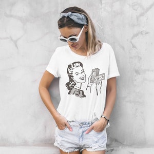 May include: White t-shirt with a vintage illustration of a woman holding a stack of money. The woman is smiling and looking at the money. The text on the money is "$100".