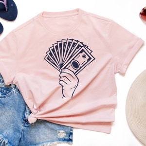 May include: A pink t-shirt with a graphic of a hand holding a stack of bills. The graphic is in navy blue.