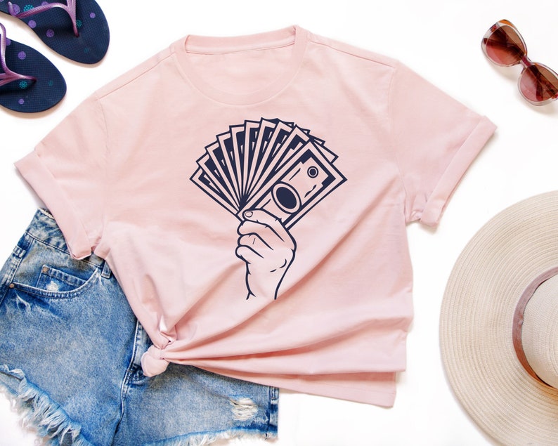 May include: A pink t-shirt with a graphic of a hand holding a stack of bills. The graphic is in navy blue.