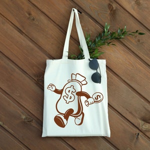 May include: White tote bag with a brown cartoon illustration of a money bag with a dollar sign and a top hat. The money bag is holding a smaller money bag with a dollar sign. The bag is on a wood background.