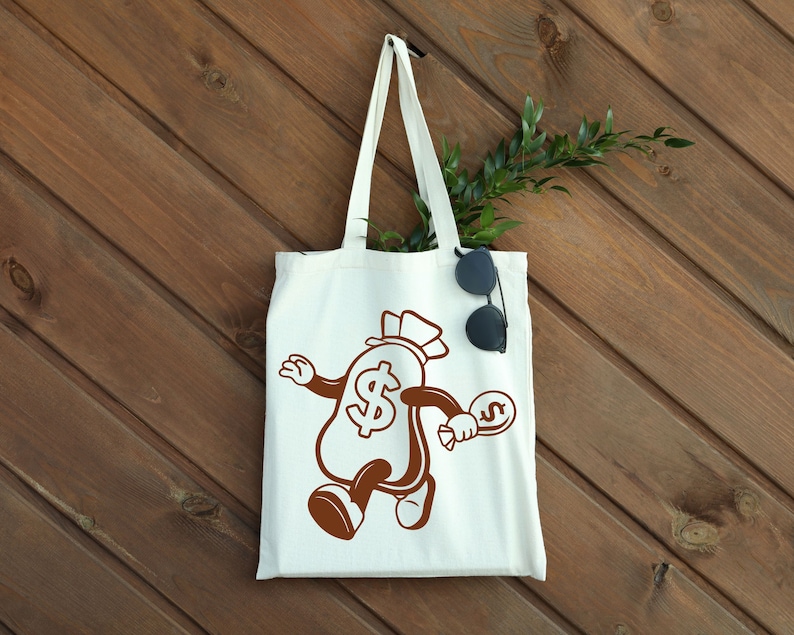 May include: White tote bag with a brown cartoon illustration of a money bag with a dollar sign and a top hat. The money bag is holding a smaller money bag with a dollar sign. The bag is on a wood background.