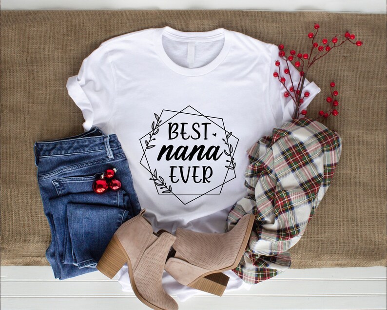 May include: White t-shirt with a black graphic that says "BEST nana EVER" in a geometric frame with floral accents. The shirt is laid out on a brown burlap surface with blue jeans, brown boots, and a plaid scarf.