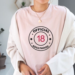 以下が含まれることがあります： 黒と白の円形のグラフィックが描かれたピンクのTシャツ。白文字で「OFFICIAL 18 ADULTHOOD」と書いてあり、赤い数字の18が書かれている。