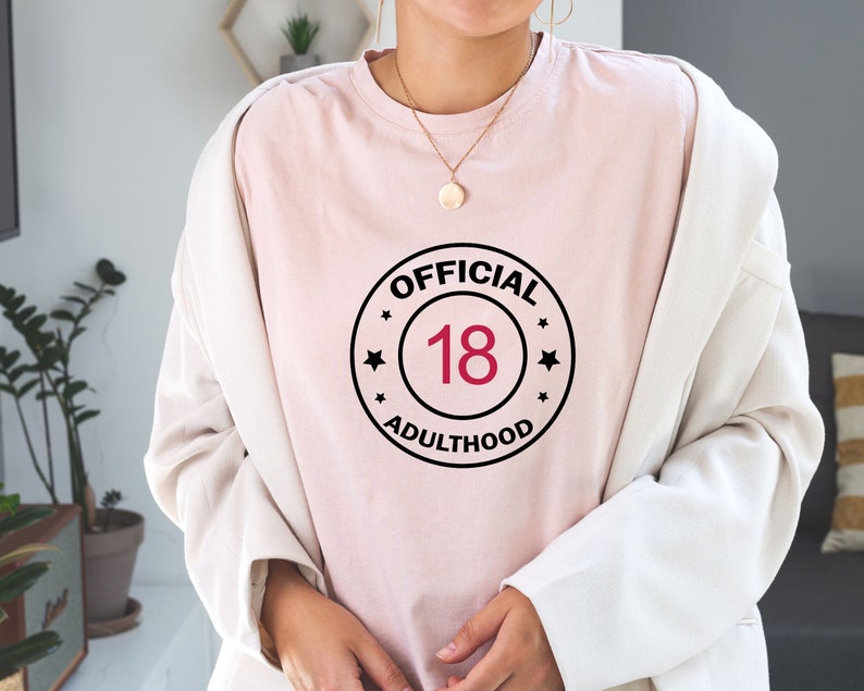 以下が含まれることがあります： 黒と白の円形のグラフィックが描かれたピンクのTシャツ。白文字で「OFFICIAL 18 ADULTHOOD」と書いてあり、赤い数字の18が書かれている。