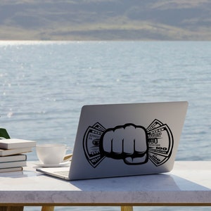 May include: A silver laptop computer with a black fist holding dollar bills on the screen. The laptop is on a white table with a cup and books.
