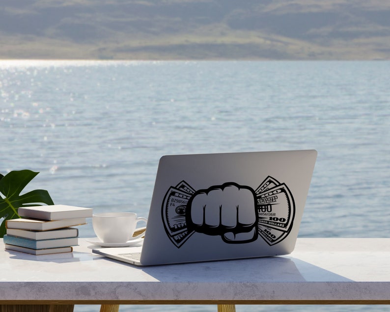May include: A silver laptop computer with a black fist holding dollar bills on the screen. The laptop is on a white table with a cup and books.