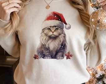 May include: A cream-colored sweatshirt featuring a watercolor illustration of a fluffy gray cat wearing a Santa hat. The cat is surrounded by snowflakes and red autumn leaves. The text "300 DPI PNG FILE" is printed at the bottom.