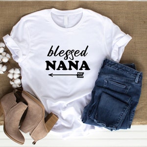 May include: White t-shirt with the text "blessed NANA" and an arrow pointing to the right. The shirt is laid out on a brown burlap surface with a pair of brown suede ankle boots and blue jeans.