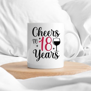 以下が含まれることがあります： 白いセラミック製のマグカップに、黒い文字で「Cheers to 18 Years」と書いてあり、18は赤色で、ワイングラスの黒いシルエットが描かれています。