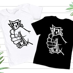 May include: Two t-shirts with a graphic of a hand holding a stack of money. The graphic is in black and white. The t-shirts are one white and one black.
