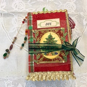 May include: A red and gold Christmas journal with a green ribbon and a beaded charm. The journal features a Christmas tree illustration and the word "JOY". The edges are trimmed with gold fringe.