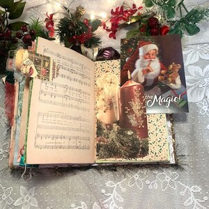 May include: Open journal with sheet music and Christmas-themed decorations. A postcard of Santa Claus holding a teddy bear is visible. The journal is decorated with greenery, berries, and candles, creating a festive holiday theme.