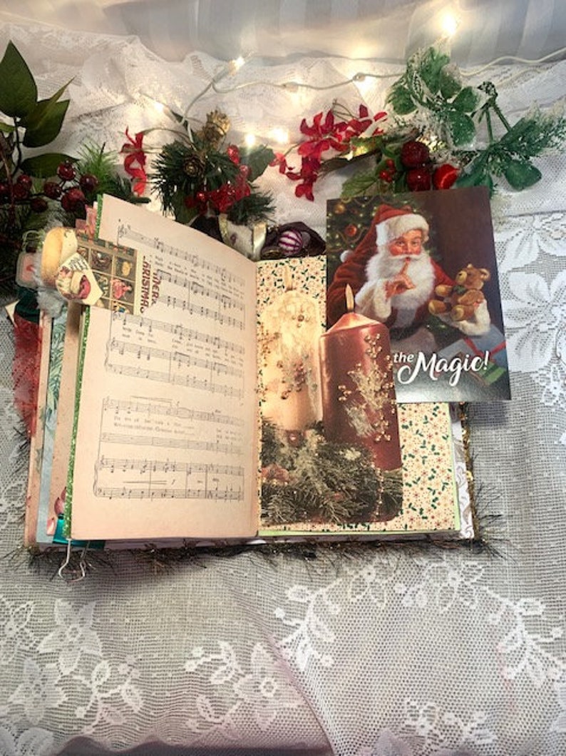 May include: Open journal with sheet music and Christmas-themed decorations. A postcard of Santa Claus holding a teddy bear is visible. The journal is decorated with greenery, berries, and candles, creating a festive holiday theme.