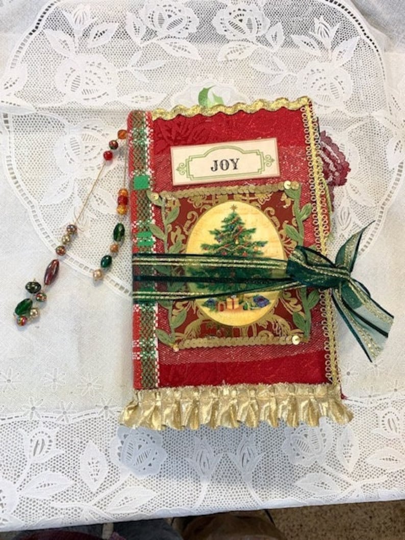 May include: A red and gold Christmas journal with a green ribbon and a beaded charm. The journal features a Christmas tree illustration and the word "JOY". The edges are trimmed with gold fringe.
