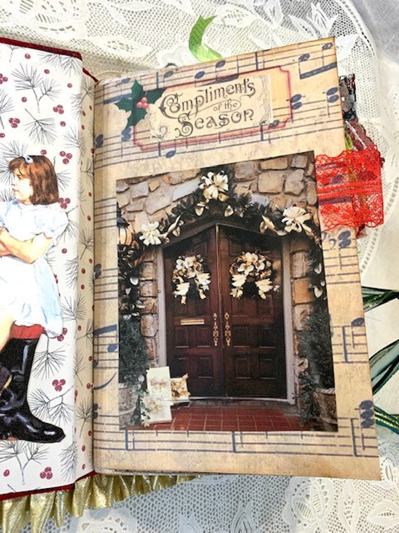 May include: Open journal with a Christmas theme. The right page features a photograph of a decorated doorway with the text "Compliments of the Season." The left page has a printed illustration of a girl in a dress and boots. The journal is trimmed with gold fabric.