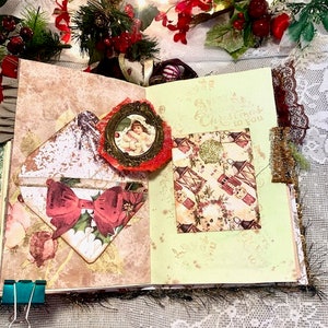 May include: Open scrapbook with Christmas-themed embellishments. Features a red bow, a small framed image of an angel, and a patterned card with a gold glitter accent. The pages have a vintage aesthetic with floral and holiday motifs.