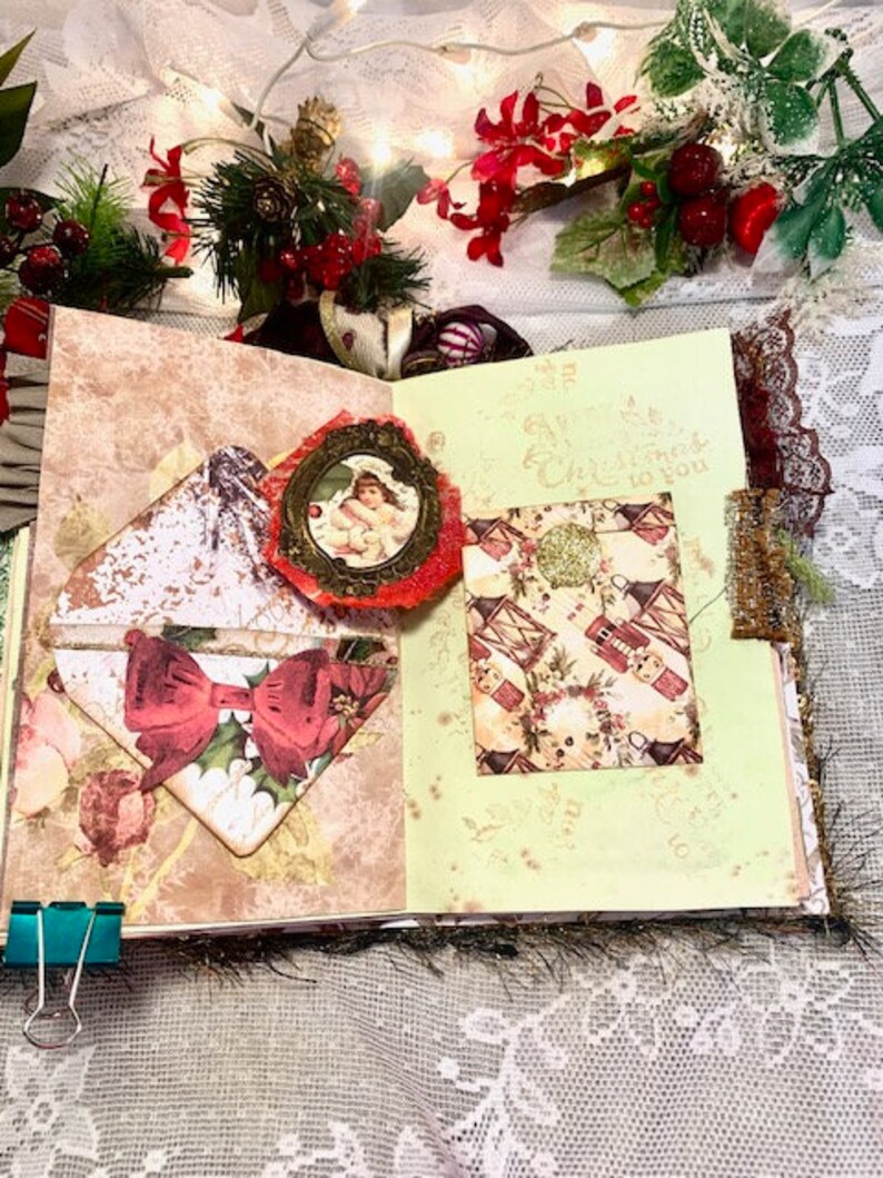 May include: Open scrapbook with Christmas-themed embellishments. Features a red bow, a small framed image of an angel, and a patterned card with a gold glitter accent. The pages have a vintage aesthetic with floral and holiday motifs.