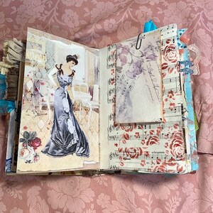 May include: Open journal with vintage-style pages. One page features a drawing of a woman in a long gown. The opposite page has sheet music with red roses. A paper with a floral design is clipped to the journal.