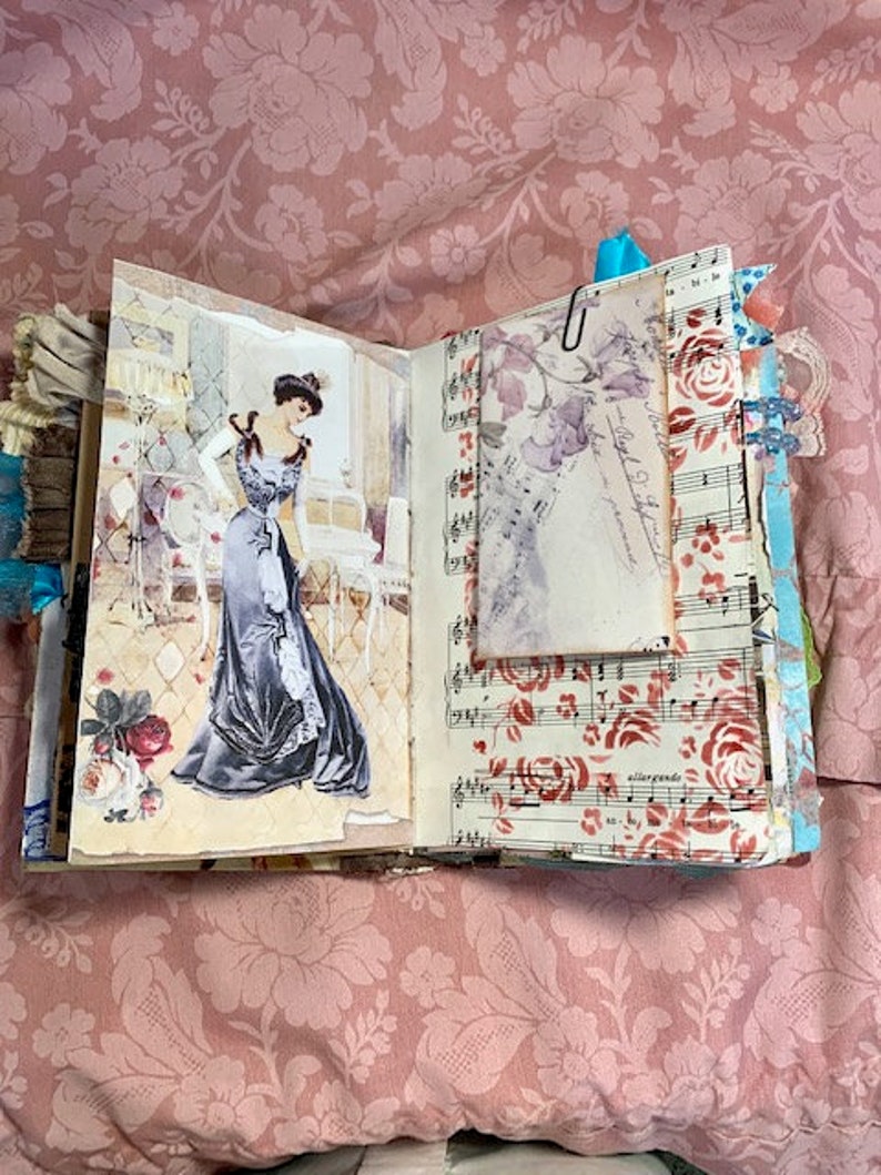 May include: Open journal with vintage-style pages. One page features a drawing of a woman in a long gown. The opposite page has sheet music with red roses. A paper with a floral design is clipped to the journal.