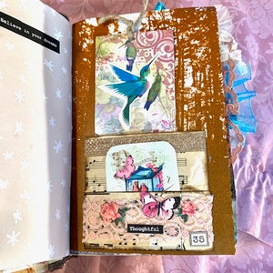 May include: Open journal with decorative pages. Features a hummingbird tag, butterfly and floral embellishments, and the word "Thoughtful". The journal's pages are adorned with musical notes and a "35" tag.