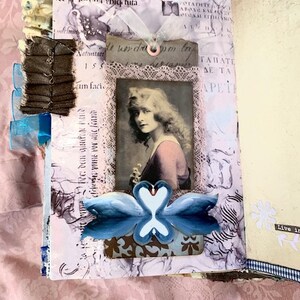 May include: A handmade journal with a vintage aesthetic. The open pages feature a portrait of a woman, lace trim, and a blue swan design. The journal is decorated with ribbons and fabric embellishments, creating a layered, artistic look.
