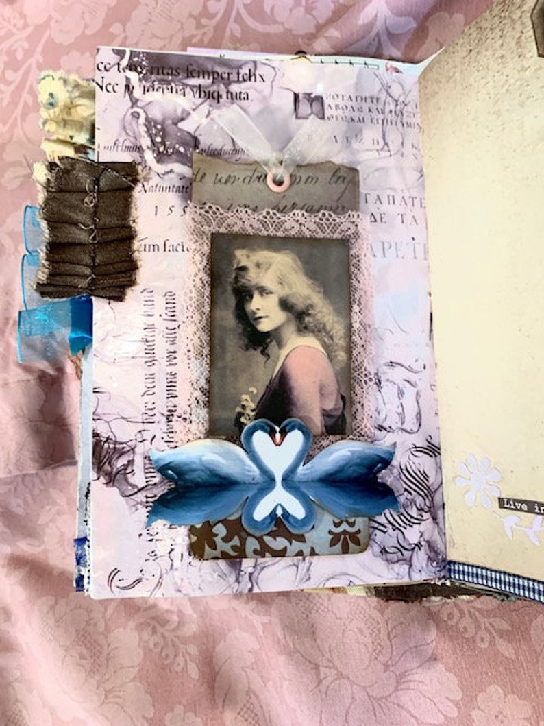 May include: A handmade journal with a vintage aesthetic. The open pages feature a portrait of a woman, lace trim, and a blue swan design. The journal is decorated with ribbons and fabric embellishments, creating a layered, artistic look.