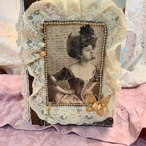 May include: A vintage-style journal with a sepia-toned portrait of a woman in a hat. The journal is adorned with cream-colored lace, gold beads, and a gold butterfly charm. The cover is framed with a beaded border.