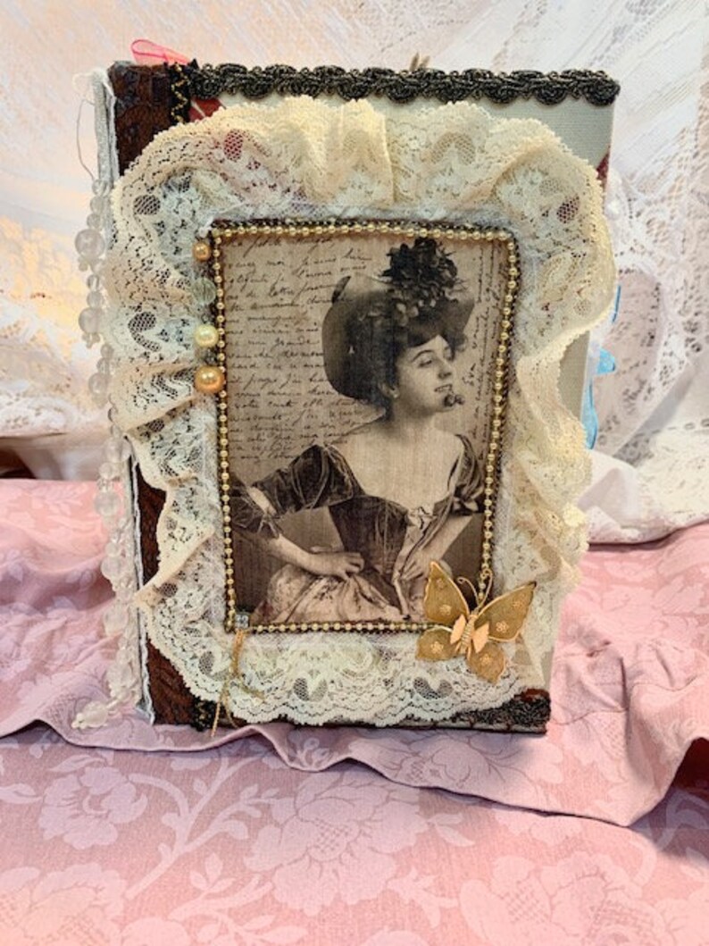 May include: A vintage-style journal with a sepia-toned portrait of a woman in a hat. The journal is adorned with cream-colored lace, gold beads, and a gold butterfly charm. The cover is framed with a beaded border.